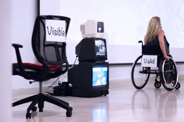 Chair with a sign reading 'visible' next to a stack of televisions and Spirit Synott in a wheelchair with the word 'valid' pinned to the back of the chair